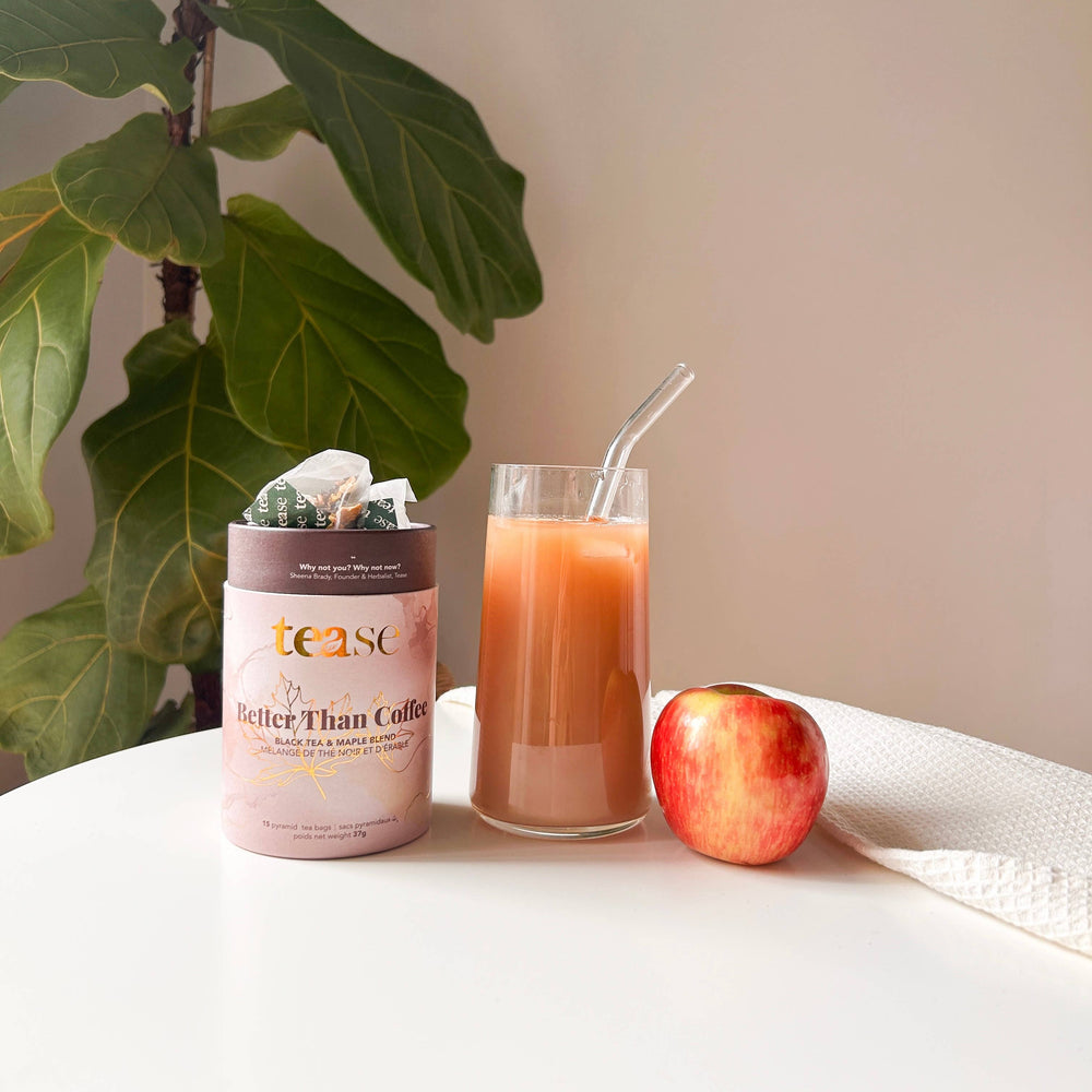 Tea canister, glass of juice, and apple on a table with a plant in the background