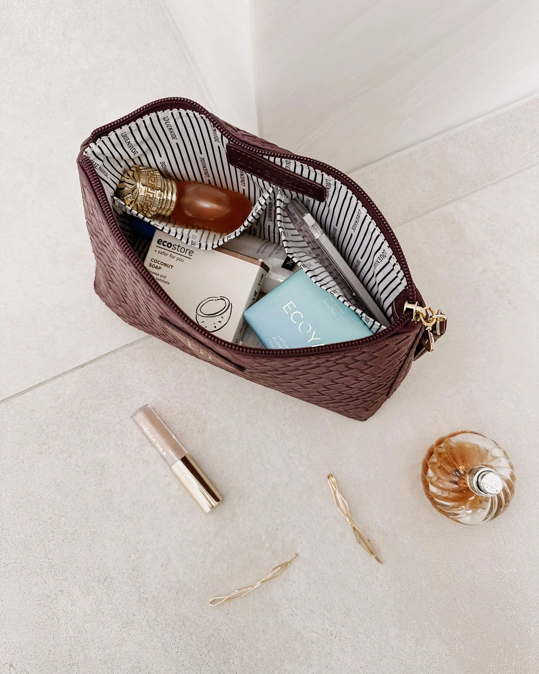 Burgundy woven makeup bag with cosmetic items on a light surface