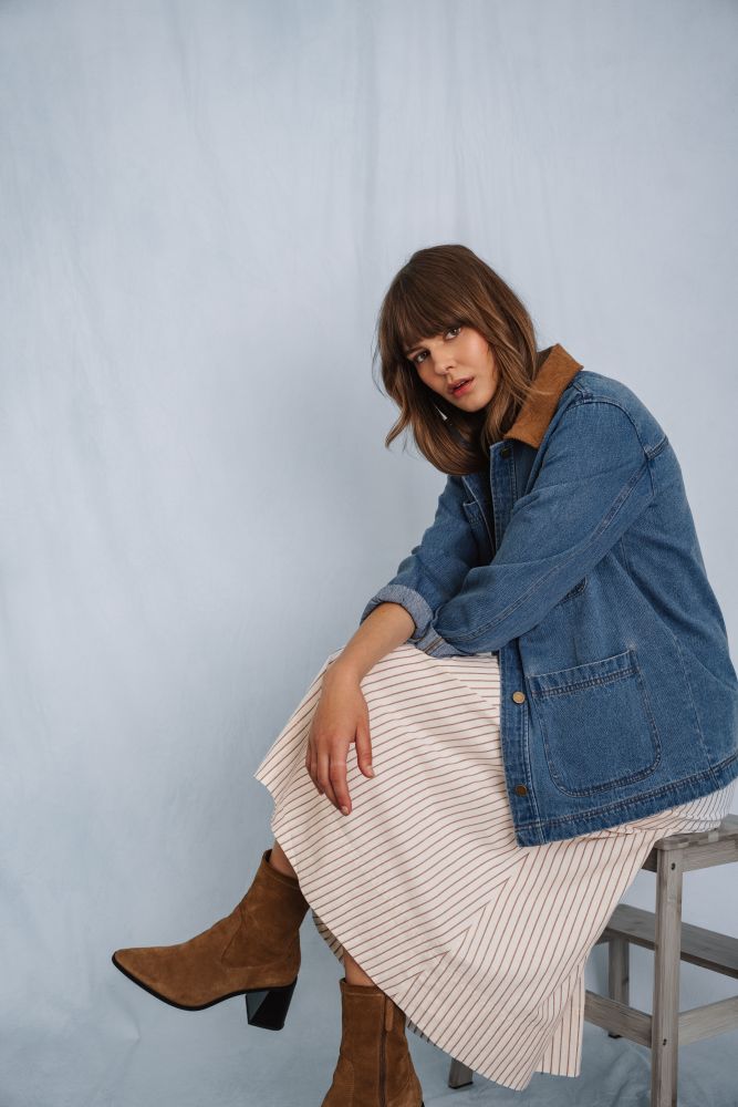 Woman wearing a denim barn jacket and beige skirt sitting on a stool against a light blue background