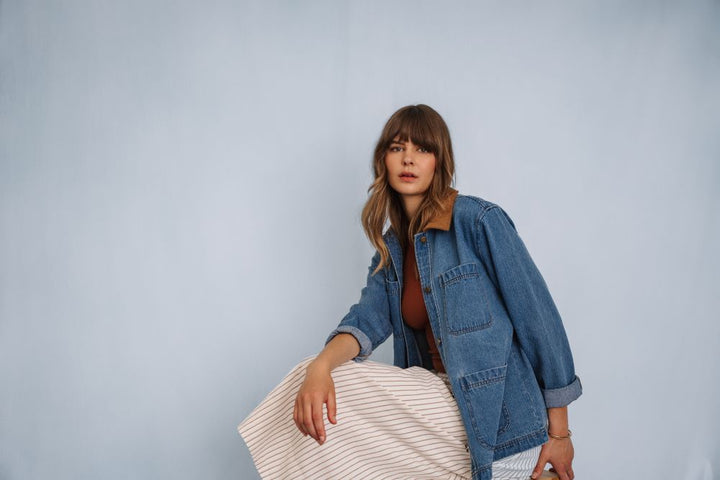 Woman wearing a denim barn  jacket holding a striped bag against a plain background