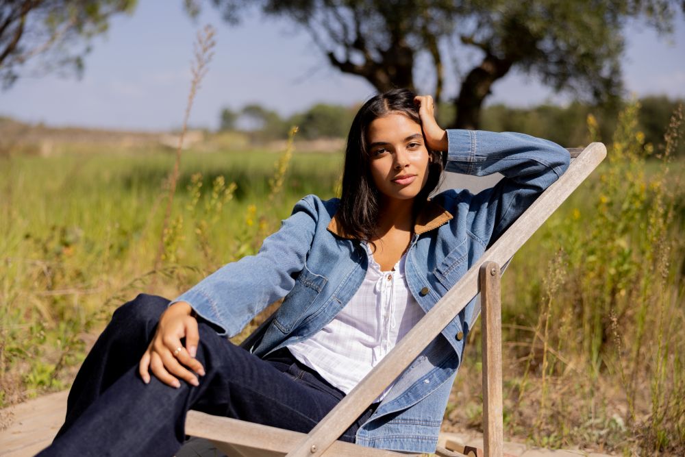 Woman wearing a denim barn jacket sitting on a wooden chair in a field with trees in the background