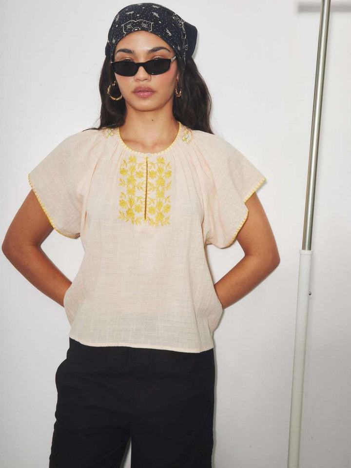 Woman wearing a beige top with yellow embroidery and black pants against a white background