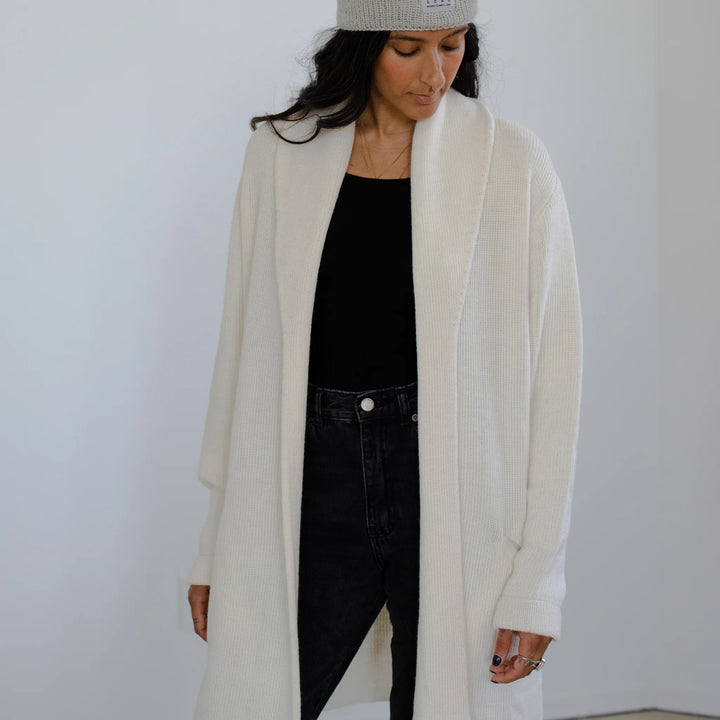Person wearing a long cream cardigan over a black outfit against a plain background