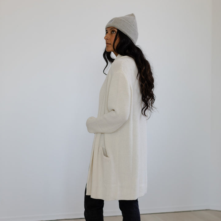 Woman wearing a long cream coat and gray beanie against a white background