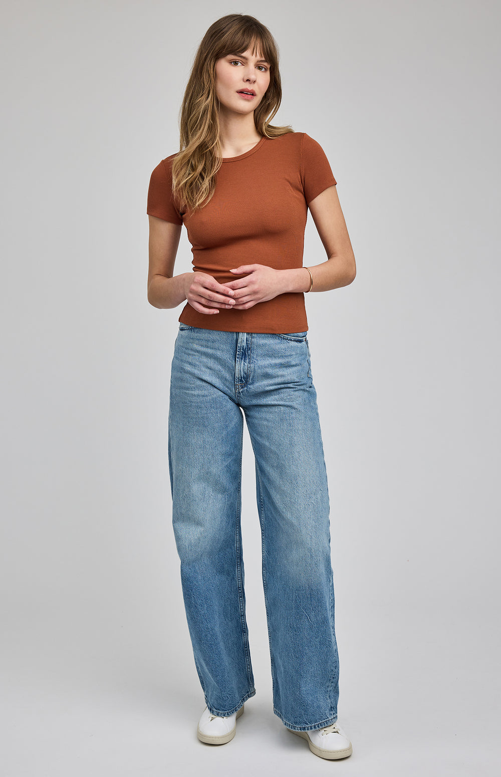 Woman wearing a rust brown ribbed t shirt and blue jeans on a gray background