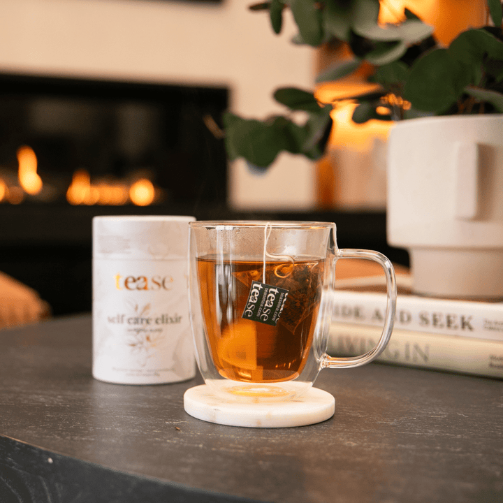 Clear mug with Self Care Herbal Tea on a marble coaster next to a white container labeled 'tease' in a cozy setting with a fireplace.
