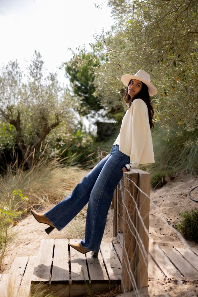 Woman in a white sweater and blue jeans sitting on a wooden bridge with trees in the background