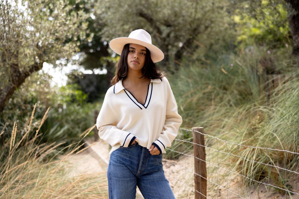 Woman wearing a white sweater with black trim and a wide-brimmed hat standing in a natural setting.