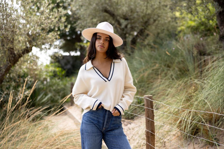 Woman wearing a white sweater with black trim and a wide-brimmed hat standing in a natural setting.