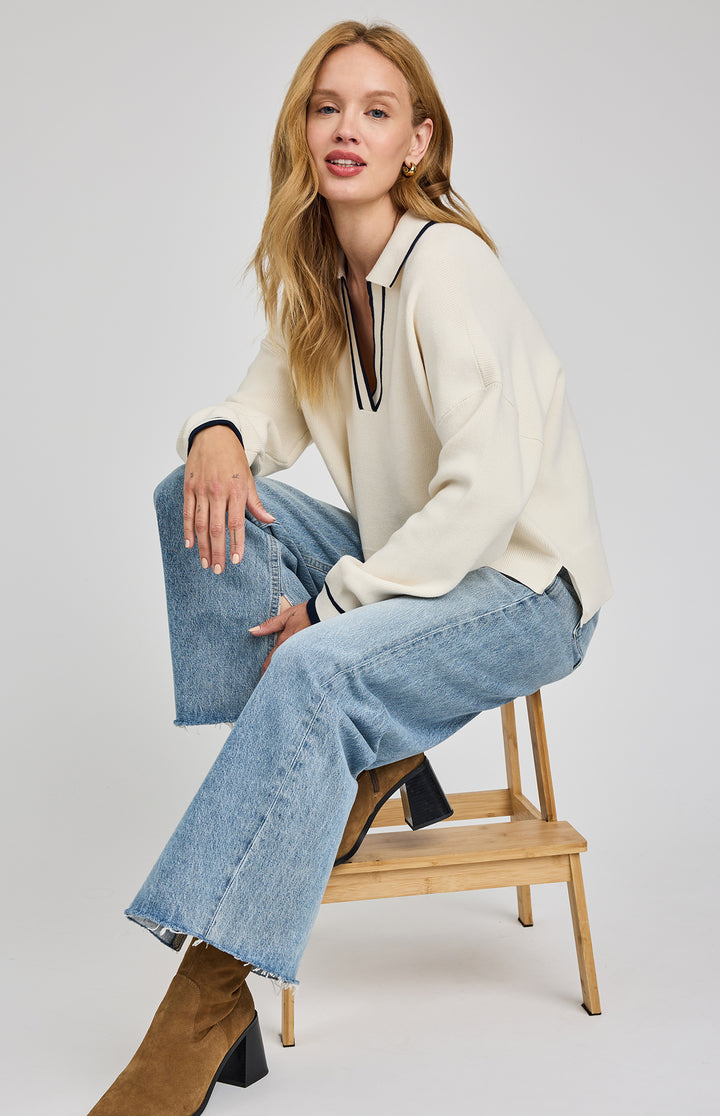 Woman sitting on a wooden stool wearing a white sweater, light blue jeans, and brown boots.