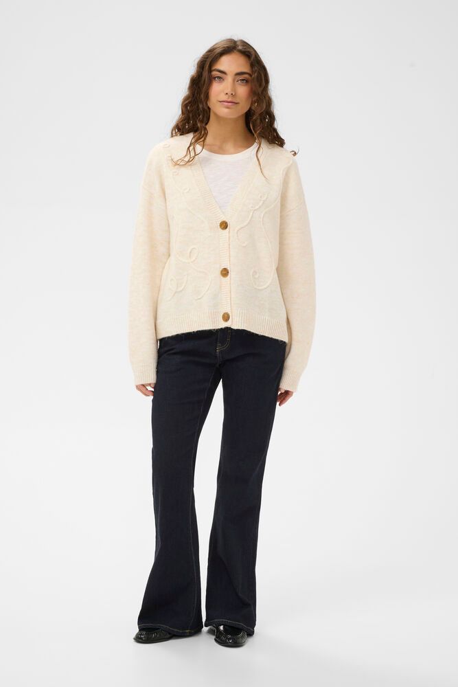 Woman wearing a cream cardigan with gold buttons, white shirt, and black pants on a white background