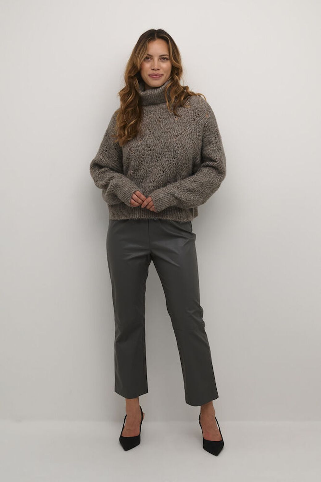 Woman wearing a  taupe brown turtleneck sweater and gray pants against a white background