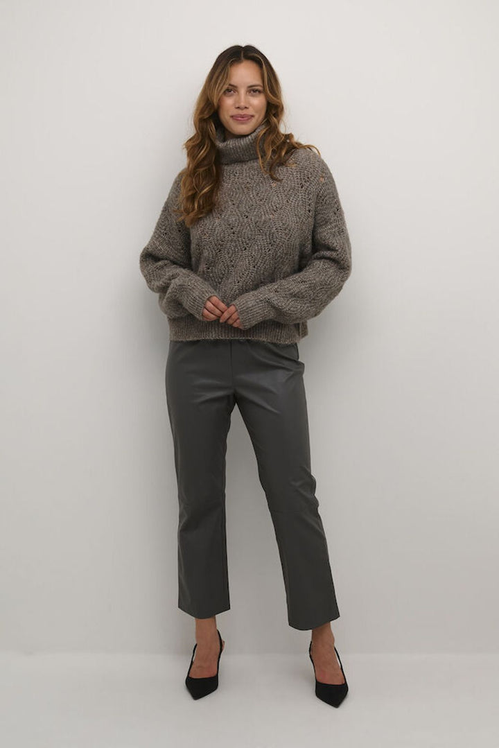Woman wearing a  taupe brown turtleneck sweater and gray pants against a white background