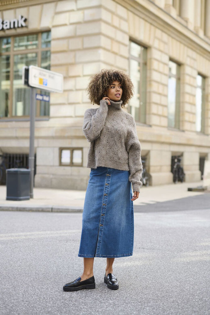 Woman wearing a taupe brown sweater and blue denim skirt standing on a street with a building in the background.