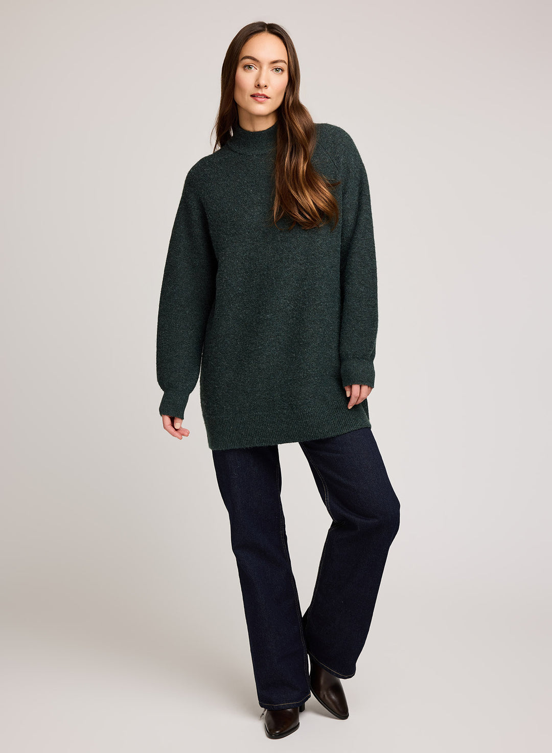 Woman wearing a green knit tunic sweater and dark pants on a plain background