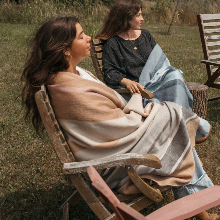 Woman sitting outdoors on a wooden chair with an alpaca wool blanket with beige and grey striped pattern  draped over her.