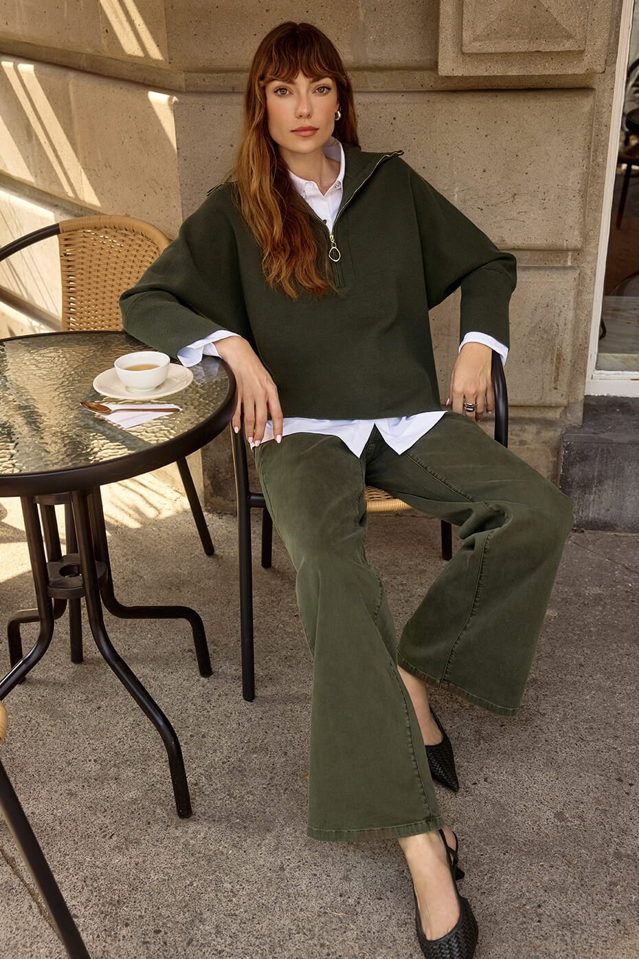 Woman sitting at an outdoor cafe table wearing a green sweater and green pants.