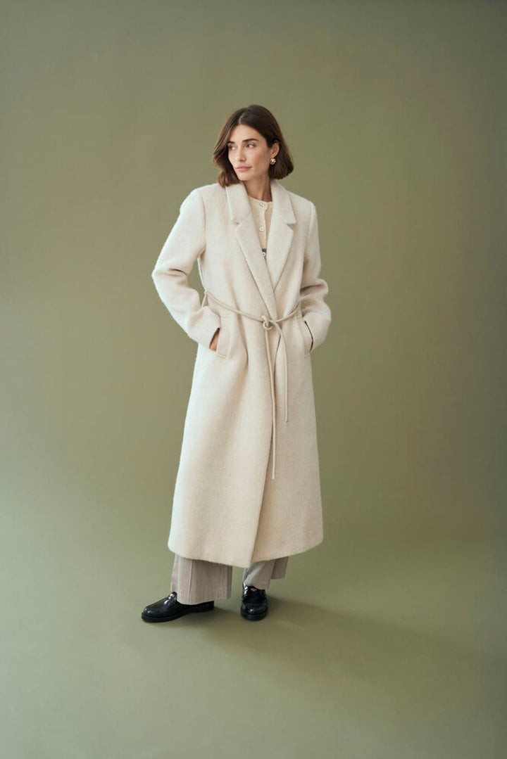 Person wearing a long cream coat against a plain background